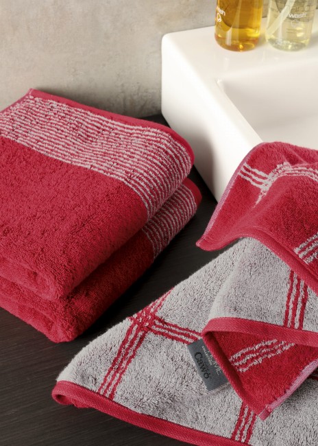 Ręcznik bawełniany 80x150 w kolorze krwistej czerwieni z szarym wzorem kraty, leżący na ciemnym blacie. W zasięgu wzroku znajduje się logo na etykiecie oraz dwa butelki z kosmetykami. W tle widoczna szara ściana.