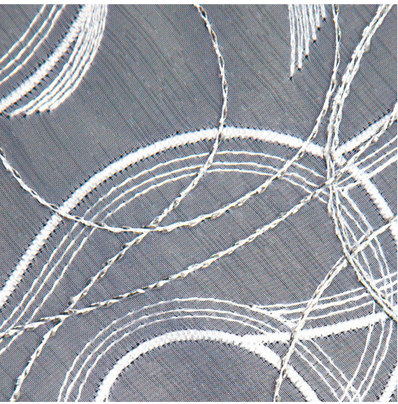 Biała firana haftowana o wysokości 280 cm, z grafitowym tłem. Zbliżenie na detal haftu przedstawiającego geometryczne wzory w postaci falistych linii, wykonanych białą nicią, dodających tekstury i elegancji.