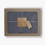 Pościel bawełniana w kolorze szaro-naturalnym, rozmiar 220x200, z falbanką na brzegach. Produkt zapakowany w eleganckie pudełko z okienkiem, przewiązane naturalnym sznurkiem. Widoczne etykiety z logo i informacjami o marce.
