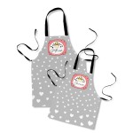 Dwa fartuchy kuchenne w szarym kolorze z białymi sercami. Fartuch dla mamy z napisem Królową Mama oraz fartuch dla córki z napisem Królową Córcia. Ujęcie frontalne, fartuchy wiszące na czarnych ramiączkach.