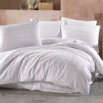 Pościel satyna bawełniana 140x200 w kolorze białym, z wzorem w delikatne kratki. Złożona, widoczne poduszki w różnych rozmiarach. Tło w odcieniach beżu, z dekoracjami w tle. Produkt w zestawieniu do użytku w sypialni.