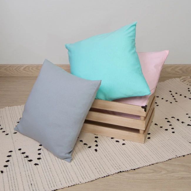 Poszewka jersey 40x40 w kolorze chabrowym, leżąca na plecionym dywaniku. W tle widoczna drewniana skrzynka z poszewkami w pastelowych kolorach: błękitnej, różowej i szarej. Produkt ma prostokątny kształt i jest rozłożony.