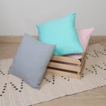 Poszewka jersey w kolorze czerwonym o wymiarach 40x40 cm, leżąca na podłodze obok drewnianego koszyka, w otoczeniu innych poszewek w pastelowych kolorach. Tło jasne, z tekstylnym, wzorzystym dywanikiem.