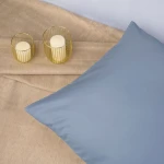 Poszewka satynowa 50x60 w kolorze niebieskim, leżąca na jasnym, teksturowanym tle. Widoczny gładki materiał, bez wzorów. W tle dwie okrągłe świeczki w metalowych osłonkach. Ujęcie z góry, ukazujące główny element w centralnej części kadru.
