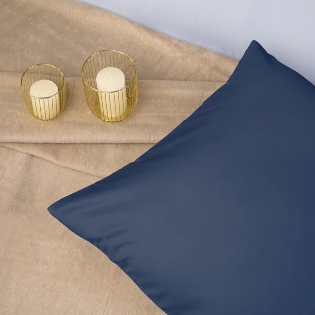 Poszewka satynowa 50x60 w kolorze chabrowym, leżąca na beżowym tle. Widoczny gładki materiał, bez wzorów. Obok dwa świeczniki w złotych metalowych osłonach z białymi świecami. Ujęcie pod kątem, eksponujące zarówno poszewkę, jak i otoczenie.