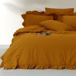 Pościel bawełniana z falbanką 220x200 Stonewashed Exclusive w kolorze musztardowym, rozłożona na łóżku. Widoczne dwie poduszki z falbanką oraz narzuta z delikatnym marszczeniem.