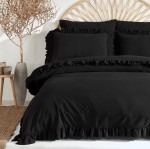 Pościel bawełniana z falbanką w kolorze czarnym, w rozmiarze 160x200 cm. Złożona na łóżku, z dekoracyjnymi falbankami przy poszewkach. W tle pleciona głowa wezgłowia i roślinność w doniczce. Tło jasne, co podkreśla produkt.