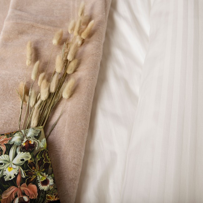 Pościel satyna bawełniana 220x200 w kolorze kremowym, ukazująca delikatny wzór w pionowe paski. Rozłożona na łóżku obok jasnego koca. Dodatkowo widoczny bukiet suszonych kwiatów w kolorze beżowym, wkomponowany w aranżację.