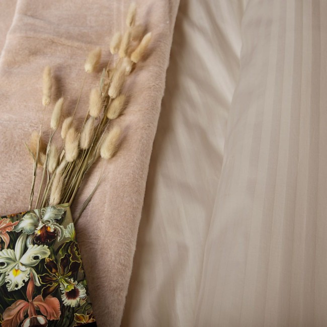 Pościel satyna bawełniana 220x200 w odcieniu jasnego beżu z delikatnym prążkiem, leżąca na podłożu. Obok widoczny bukiet suchego kwiatu w tonalnych beżach oraz kwiatowa okładka z intensywnym nadrukiem, tworząca stonowaną aranżację.