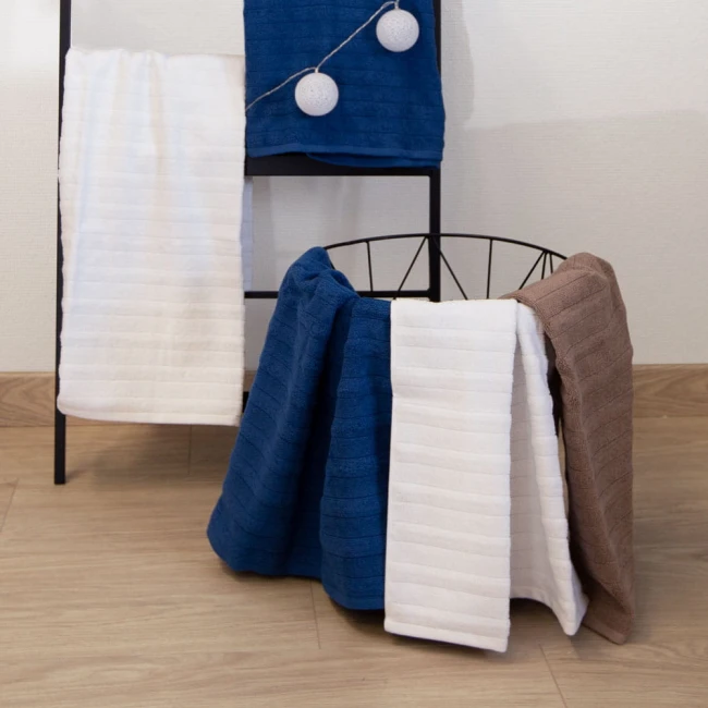 Ręcznik bawełniany w kolorze brązowym o wymiarach 50x90 cm, z widoczną teksturą i poziomymi paskami. Leży wyłożony na metalowej półce, obok ręczników w kolorze niebieskim i białym. Tło jest jasne, co podkreśla kolorystykę produktów.