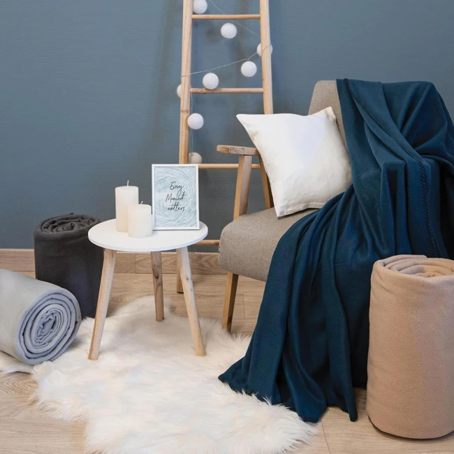 Koc polarowy 150x200 cm w kolorze butelkowej zieleni, rozłożony na szarej sofie obok białej poduszki. W tle stojak z lampkami i zielona ściana. Obok znajdują się dwa zrolowane koce w kolorach szarym i beżowym, a na stoliku trzy świece.