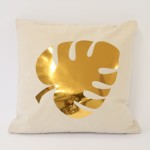 Poszewka dekoracyjna 40x40 cm w kolorze jasnym z połyskującym, złotym wzorem liścia monstera. Produkt leży na jednolitym tle. Materiał jest gładki, z miękką fakturą. Wzór jest wyraźny, bez dodatkowych napisów czy logo.