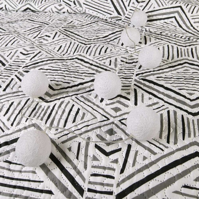 Narzuta drukowana Tarik 170x210 w biało-czarnym geometrycznym wzorze, leżąca na jasnym tle. Widoczne dekoracyjne kulki z białego materiału, rozłożone wzdłuż narzuty, dodające efektu stylowego wystroju. Idealna do nowoczesnych aranżacji wnętrz.