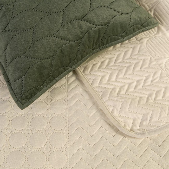 Narzuta Verde mink w kolorze zielonym o wymiarach 170x210 cm, z pikowaniem w wzory w kształcie fal i rombów. Zdjęcie przedstawia narzutę leżącą na podłożu, z widoczną poduszka w kolorze zielonym z pikowaniem.