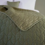 Narzuta Verde mink w kolorze zielonym, o wymiarach 240x220 cm, z delikatnym, falistym wzorem i matową fakturą. Ułożona na łóżku, z widocznym poduszką w tym samym kolorze, prezentuje elegancki styl.