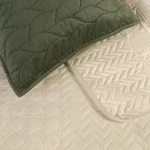 Narzuta Verde mink 240x220 w kolorze zielonym z pikowanym wzorem w kształcie fal. Na zdjęciu widoczna narzuta na jasnym tle z poduszką w kolorze zielonym, z różnymi fakturami: okrągłymi i zygzakowymi.