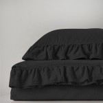 Pościel bawełniana z falbanką w kolorze czarnym, złożona na łóżku. Widoczny zestaw składający się z poszwy na kołdrę i dwóch poduszek, pokryty delikatną falbanką. Materiał o lekko zmiętej strukturze. Tło jasne, jednolite.