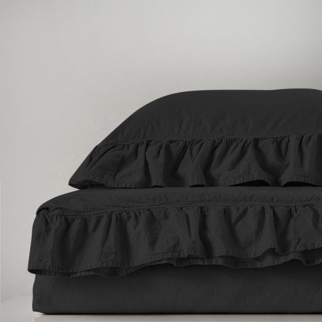 Pościel bawełniana z falbanką w kolorze czarnym, złożona na łóżku. Widoczny zestaw składający się z poszwy na kołdrę i dwóch poduszek, pokryty delikatną falbanką. Materiał o lekko zmiętej strukturze. Tło jasne, jednolite.