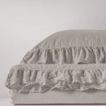 Pościel bawełniana z falbanką 220x200 w odcieniu naturalnej kremowej, leżąca na gładkiej powierzchni. Widoczny detal falbanki oraz tekstura materiału. Produkt rozłożony, ukazujący miękkość i elegancki fason. Tło jasne, jednolite.