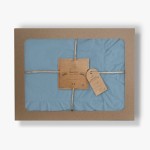 Pościel bawełniana z falbanką w kolorze błękitnym, opakowana w prostokątne pudełko z brązowego kartonu. Widoczna etykieta z logo „LIMASSO”, sznurek jako zamknięcie, detal falbany widoczny u dołu narzuty. Wymiary 160x200.