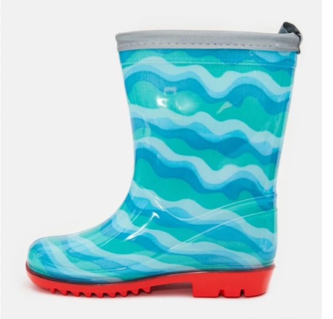Kalosze dziecięce Cool Kids w kolorze błękitnym z wzorem fal na cholewce, z czerwoną podeszwą. Ujęcie z boku, stojące, ukazujące wysoką cholewkę oraz szary brzeg. Gumowy materiał, gładka tekstura, idealne na deszczowe dni.