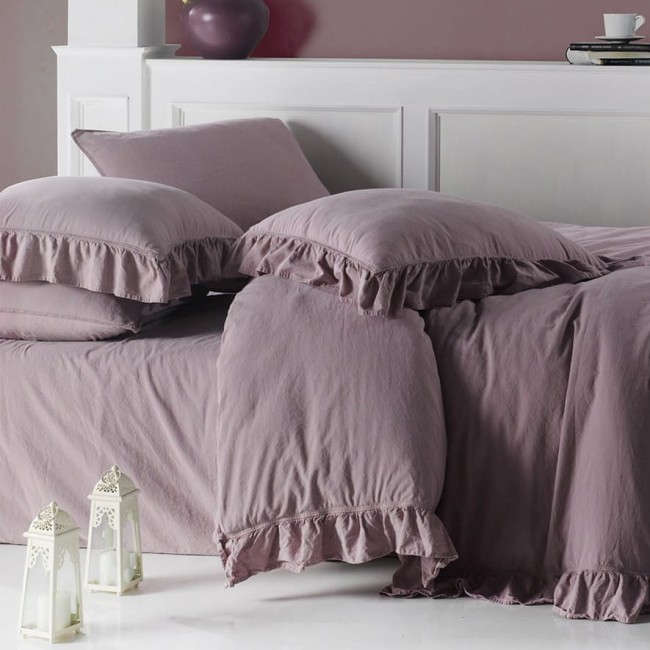 Komplet dwóch poszewek bawełnianych 40x40 cm w odcieniu fioletowym, z dekoracyjną falbanką. Ułożone na łóżku, z widoczną teksturą materiału. W tle biały parawan i subtelne detale aranżacyjne, takie jak lampiony.