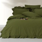 Komplet poszewek bawełnianych z falbanką w kolorze zielonym Stonewashed Exclusive Olive, rozmiar 2x40x40 cm. Ułożone na łóżku z falbanką wzdłuż krawędzi, złożone w różne pozycje. W tle jasna powierzchnia, obok gazetka.