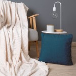 Poszewka żakardowa 40x40 w kolorze błękitu marokańskiego, z teksturowanym wzorem. Ułożona na szarej poduszce obok jasnego koca, na białym stoliku znajdują się okrągły pojemnik oraz lampka z przezroczystą żarówką. Tło w odcieniu ciemnoszarym.