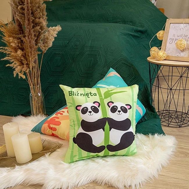 Poduszka przytulanka Bliźnięta 40x40 z ilustracją dwóch pand w odcieniach zieleni i czerni. Ułożona na miękkim dywanie, otoczona innymi poduszkami i świecami. Tło w kolorze butelkowej zieleni. W widoku frontalnym, z wyraźnym napisem Bliźnięta.