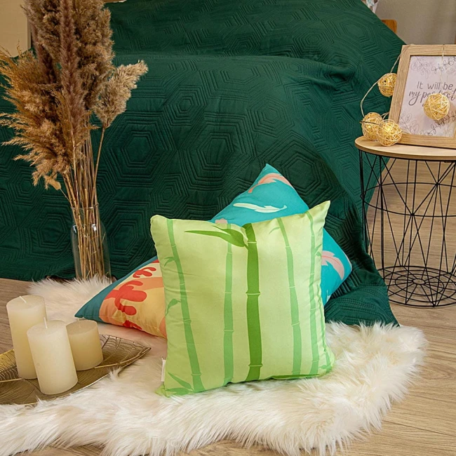 Poduszka przytulanka Bliźnięta 40x40 w zielonym kolorze z grafiką bambusa, leży na białym futerku obok poduszek w kolorach błękitu i czerwieni. W tle zielona kołdra, a obok mały stół z dekoracyjnymi świecami i lampkami.