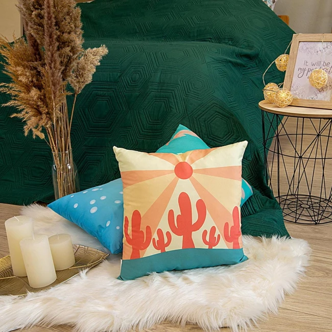 Poduszka przytulanka Koziorożec 40x40 o pastelowych kolorach w odcieniach żółtym i turkusowym z graficznym wzorem przedstawiającym kaktusy na tle słońca. Ułożona na białym, puchatym dywaniku, z otoczeniem dekoracyjnymi świecami i roślinami.