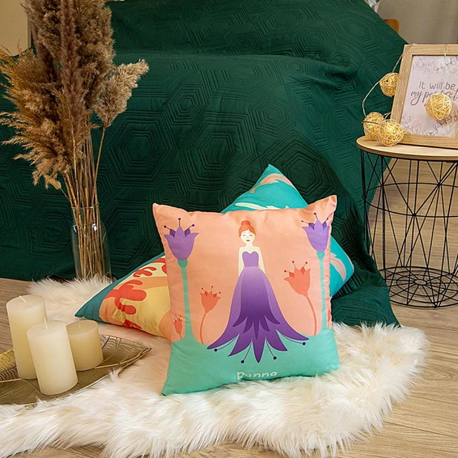 Poduszka przytulanka Panna 40x40 w odcieniach brzoskwiniowym i miętowym, z ilustracją dziewczynki w fioletowej sukience otoczonej kwiatami. Produkty ustawione leżąco na białym futerku, w tle zielona narzuta i dekoracyjna taca z świecami.