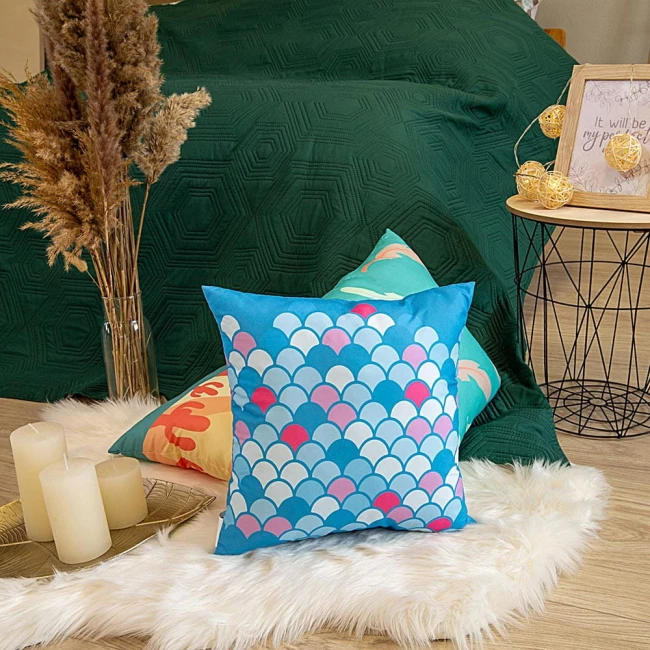 Poduszka przytulanka w kształcie ryby, rozmiar 40x40 cm, w kolorze błękitnym z wzorem w białe, różowe i niebieskie łuski. Produkt leży na białym futrze, obok kilku innych poduszek. W tle zielony koc oraz dekoracyjny stół z świecami i ozdobami.