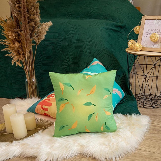 Poduszka przytulanka Waga 40x40 w kolorze zielonym, pokryta wzorem w liście w odcieniach żółtego i niebieskiego. Ułożona na podłodze na sztucznej wsypie, obok świec w metalowym świeczniku. Tło z zielonym narzutą na łóżko.