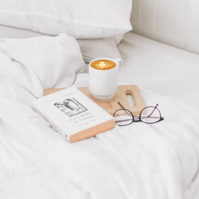 Prześcieradło bawełniane hotelowe białe 200x220, leżące na łóżku. W tle widoczne poduszki oraz kawa w białej filiżance na drewnianym stoliku, obok książka i okulary. Tło jasne i stonowane, tworzące spokojny klimat.