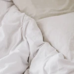 Prześcieradło bawełniane hotelowe w kolorze białym, rozłożone na łóżku, z widoczną gładką fakturą. Ułożone z boku poduszki w odcieniu beżu. Produkt w ułożeniu leżącym, na neutralnym tle. Wymiary 200x220 cm.