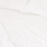 Prześcieradło bawełniane hotelowe, białe, o wymiarach 200x220 cm, rozłożone na łóżku. Gładka powierzchnia bez wzorów, prezentujące teksturę materiału. W tle widoczne poduszki o białym pokryciu.