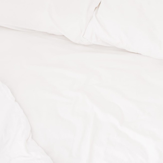 Prześcieradło bawełniane hotelowe, białe, o wymiarach 200x220 cm, rozłożone na łóżku. Gładka powierzchnia bez wzorów, prezentujące teksturę materiału. W tle widoczne poduszki o białym pokryciu.