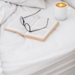 Prześcieradło bawełniane hotelowe białe 200x220 GŁADKIE na łóżku z książką i filiżanką kawy. Ujęcie z góry, produkt leży rozłożony na prostym, jasnym tle. Widoczne okulary na stronie książki.