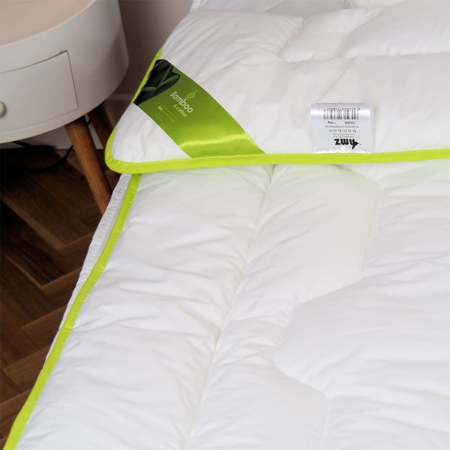 Kołdra całoroczna 200x220 cm, antyalergiczna, w kolorze białym, otoczona zieloną lamówką. Ułożona leżąco na łóżku z widoczną etykietą i logo. Tło proste, na drewnianej podłodze, co podkreśla jej kształt i detale kompozycji.