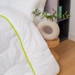 Kołdra całoroczna 200x220 cm w kolorze białym z zieloną lamówką, rozłożona na drewnianej podłodze obok dekoracyjnego okrągłego wazonu. Widoczna tekstura materiału i struktura wypełnienia. Kołdra antyalergiczna, idealna do codziennego użytku.