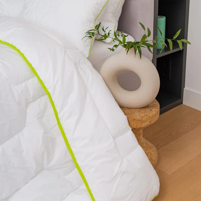 Kołdra całoroczna 200x220 cm w kolorze białym z zieloną lamówką, rozłożona na drewnianej podłodze obok dekoracyjnego okrągłego wazonu. Widoczna tekstura materiału i struktura wypełnienia. Kołdra antyalergiczna, idealna do codziennego użytku.