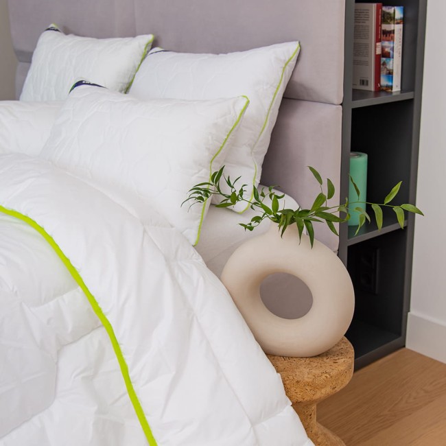 Poduszka antyalergiczna pikowana 50x60 cm w białym kolorze z zielonym obszyciem leży na pościeli. W tle widoczne trzy poduszki na wezgłowiu łóżka oraz ozdobny element w kształcie pierścienia z liśćmi na stoliku.