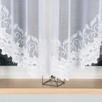 Firana żakardowa w kolorze białym o wymiarach 400x160 cm, wisząca, z białym haftowanym wzorem roślinnym na dolnej krawędzi. Tło zdjęcia jest jasne, a firana prezentuje się w stonowanej aranżacji na drewnianym parapecie.