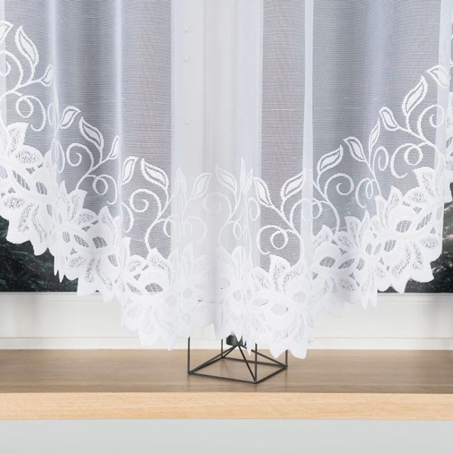 Firana żakardowa w kolorze białym o wymiarach 400x160 cm, wisząca, z białym haftowanym wzorem roślinnym na dolnej krawędzi. Tło zdjęcia jest jasne, a firana prezentuje się w stonowanej aranżacji na drewnianym parapecie.