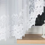 Firana gotowa Bastia żakardowa w kolorze białym, o wymiarach 400x160 cm. Niezwykle delikatny materiał z haftowanym wzorem liściowym. Produkt zwisający, widoczny z przodu, umieszczony na tle jasnego wnętrza z dekoracyjną lampionem.