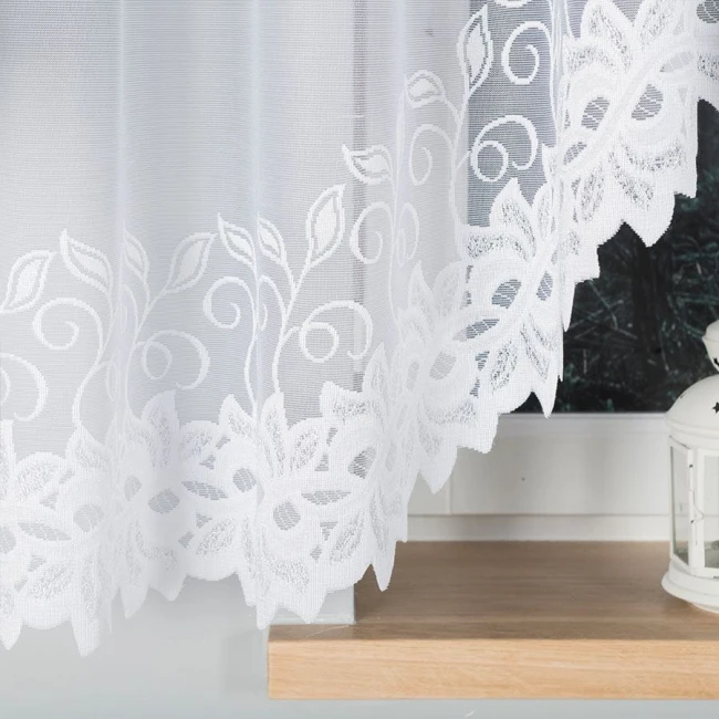 Firana gotowa Bastia żakardowa w kolorze białym, o wymiarach 400x160 cm. Niezwykle delikatny materiał z haftowanym wzorem liściowym. Produkt zwisający, widoczny z przodu, umieszczony na tle jasnego wnętrza z dekoracyjną lampionem.
