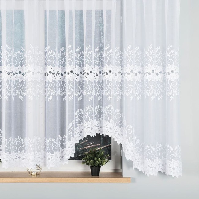Firana gotowa Bastia żakardowa biała 500x160 z delikatnym wzorem liści i kwiatów na dolnej krawędzi. Produkt zawieszony na oknie, lekko zwisający, wykonany z transparentnego materiału. Widoczna doniczka z zieloną rośliną na parapecie.