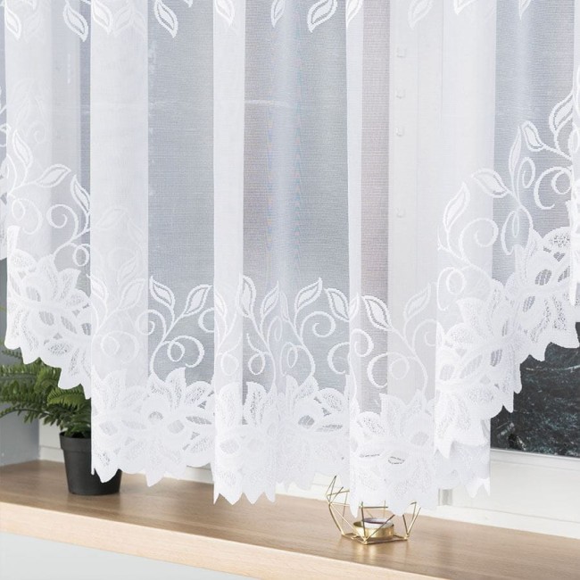 Firana gotowa Bastia, żakardowa, w kolorze białym, o wymiarach 500x160 cm. Zawiera dekoracyjny wzór roślinny na dolnej krawędzi. Produkt wiszący, częściowo przezroczysty, z delikatną teksturą, umieszczony na oknie z kwiatami na parapecie.