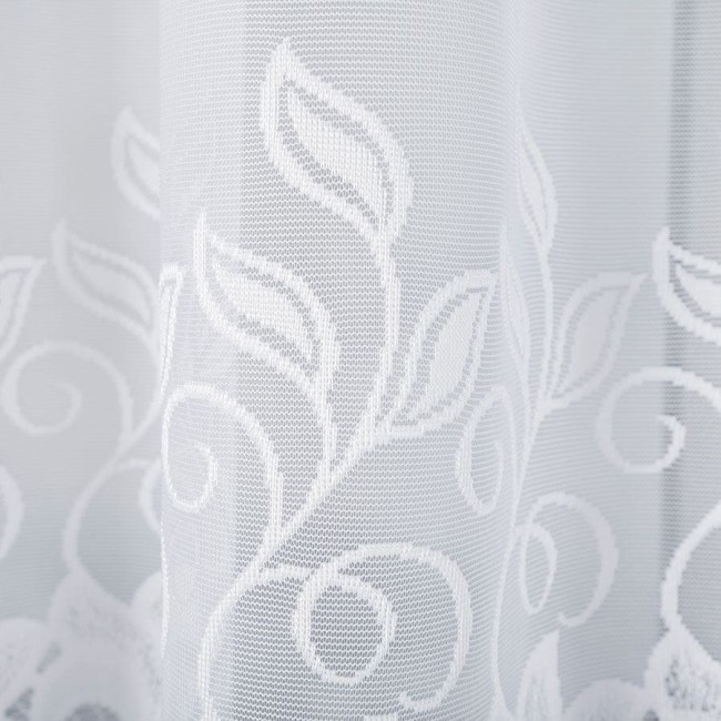 Biała firana żakardowa Bastia o wymiarach 500x160 cm, z delikatnym wzorem liści i spiral. Ujęcie zbliżeniowe, ukazujące fakturę i szczegóły wzoru. Podkreśla lekkość materiału, zawieszona, na jasnym tle.